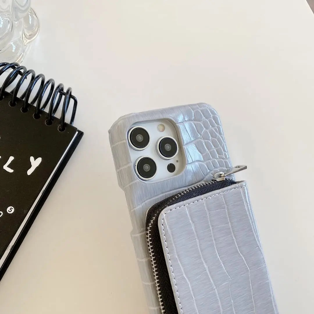 Phone case with crocodile texture and zipper on a light surface next to a black notebook.