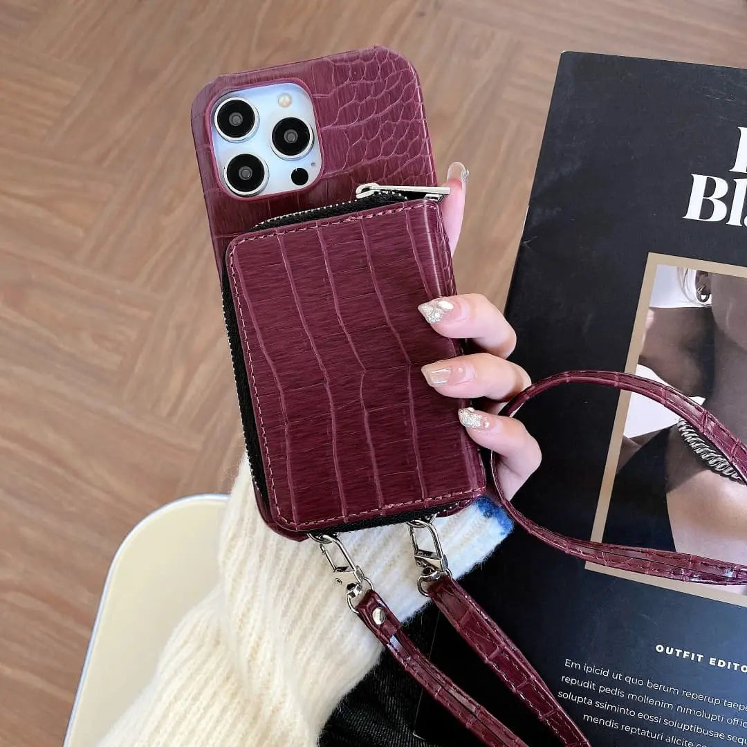 Maroon crocodile-textured wallet phone case with a strap held by a hand on a wooden surface.