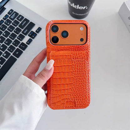 A hand holding an orange phone case with a crocodile-embossed texture, featuring a built-in card slot pocket, shown in a lifestyle setting on a white desk next to a laptop.