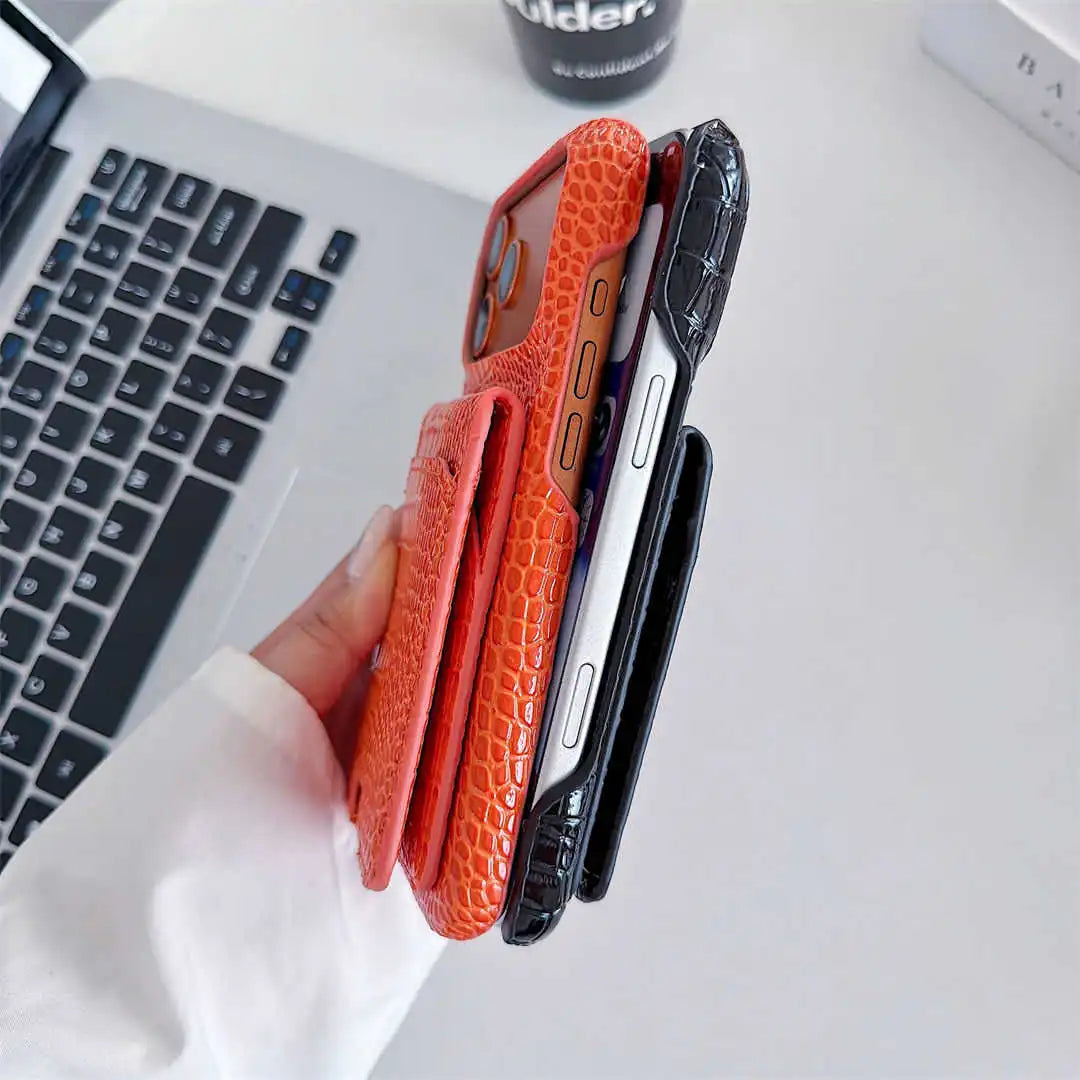 A hand holds three slim phone cases with a textured crocodile pattern in orange, black, and dark gray, shown next to a silver laptop on a white desk.