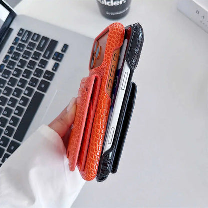 A hand holds three slim phone cases with a textured crocodile pattern in orange, black, and dark gray, shown next to a silver laptop on a white desk.