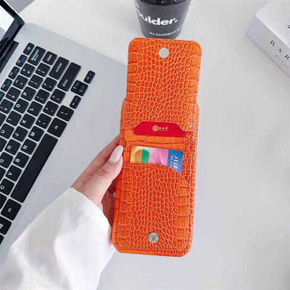 An orange crocodile-embossed leather card holder wallet held by a hand, shown open with two cards inserted. The background is a white desk with a laptop, a black cup, and a book visible.