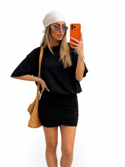 A woman takes a mirror selfie wearing a black short-sleeve t-shirt and matching black ruched mini skirt, styled with a white headscarf, sunglasses, a tan woven shoulder bag, and an orange smartphone case against a plain white background.